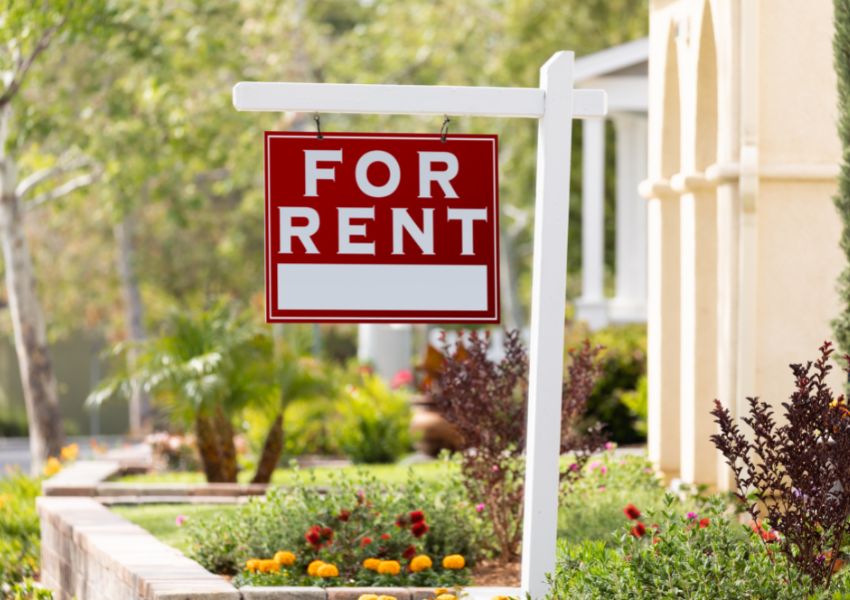A red and white 'for rent' sign hangs from a white wooden post in front of a rental property in the light of day.